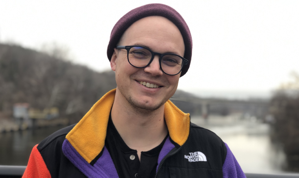 A portrait of Daniel Richtmyre. A smiling white man wearing a burgundy beanie on his head, with navy, thick-rimmed glasses. He is wearing a slightly open North Face jacket with yellow, purple, and red fabric. In the background is an out-of-focus river.